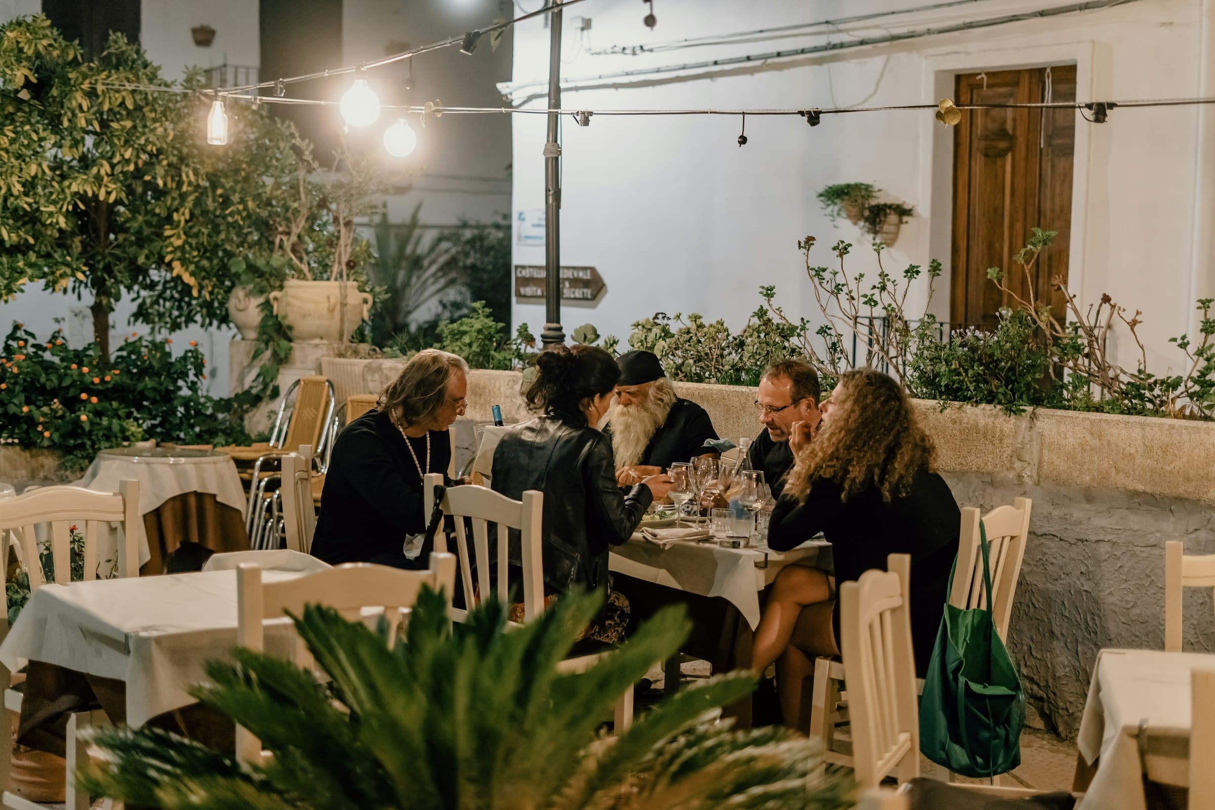 Friends gathered around a long courtyard table at dusk, talking under string lights